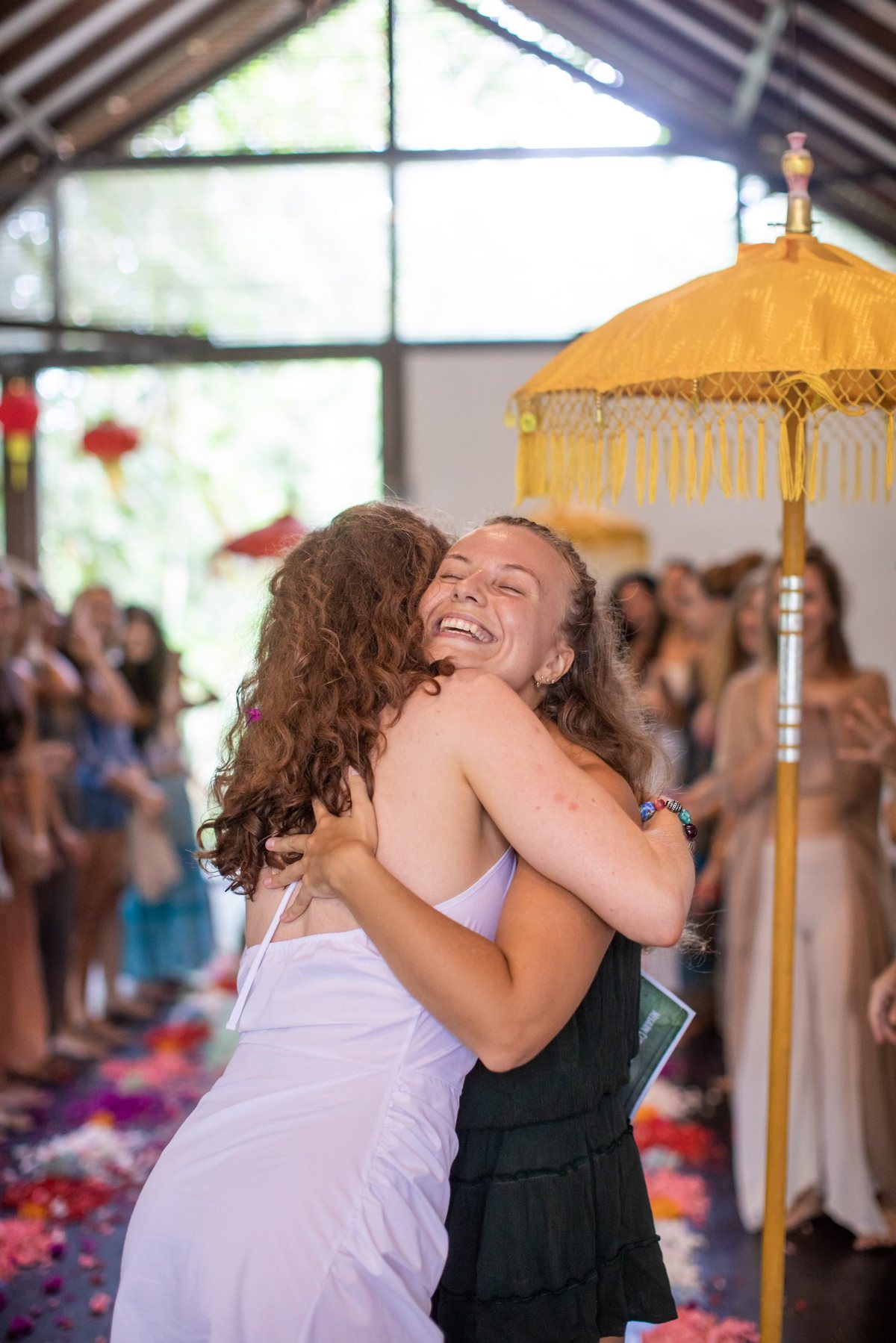 Two retreat guests hugging as they arrive, petals on the floor and a Balinese umbrella behind them
